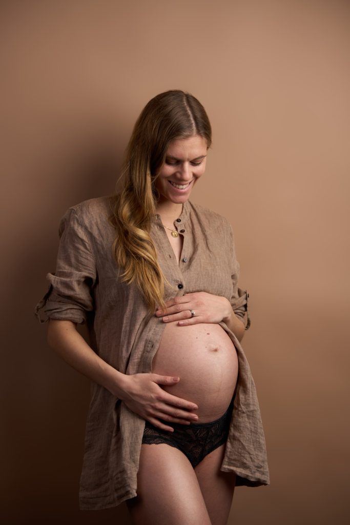 Gravid kvinde i åben brun hørskjorte og sort blonde underkjole smiler glad ned mod sin gravide mave mod terracotta baggrund