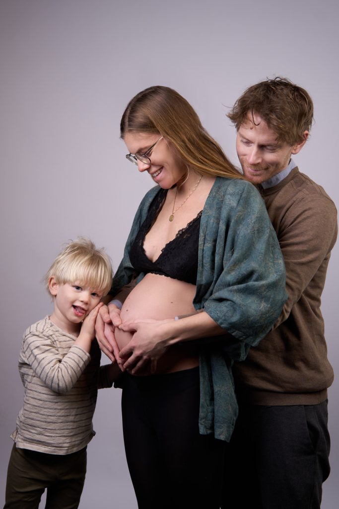 Gravid mor i teal kimono med far og lille blond dreng der alle holder hænder om den gravide mave, grå studiobaggrund