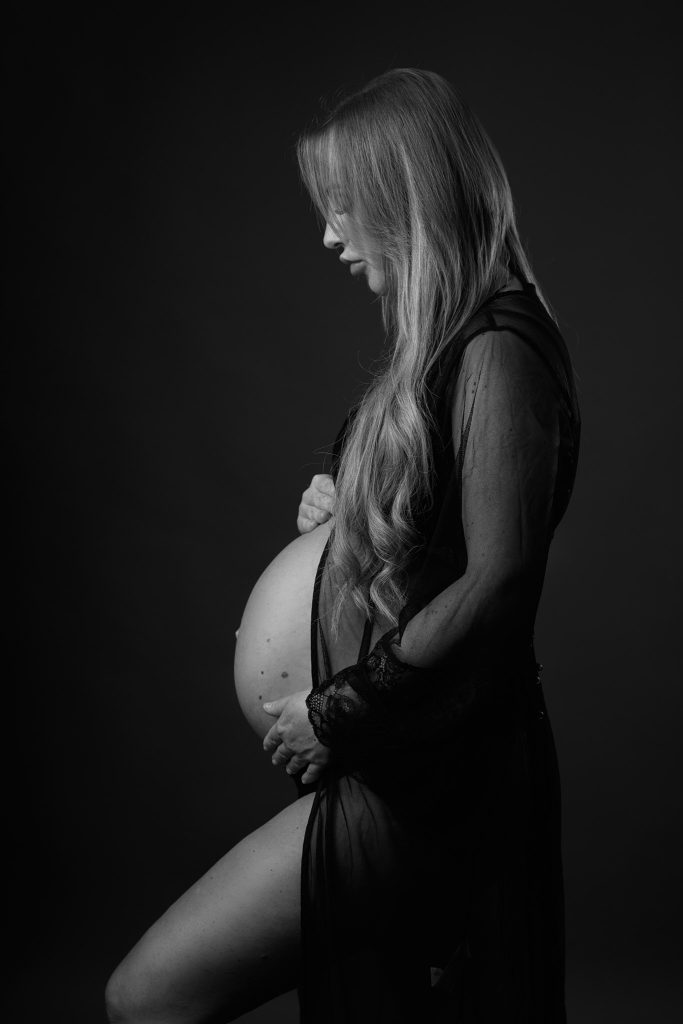 Sort-hvid graviditetsfoto af gravid kvinde i sort gennemsigtigt negligé med langt lyst hår i profil mod mørk baggrund