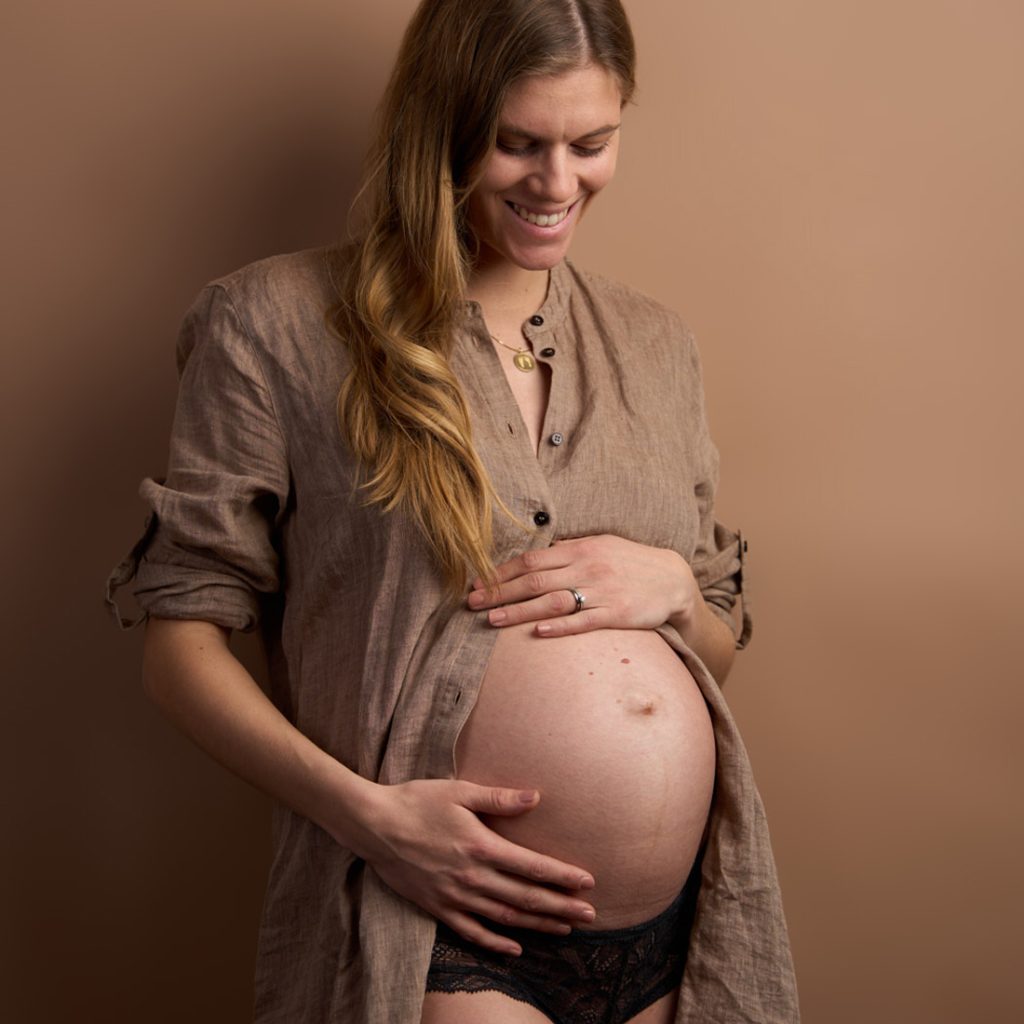 Gravid kvinde i åben brun hørskjorte og sort lingeri holder om sin store mave og smiler ned mod den, terracotta baggrund