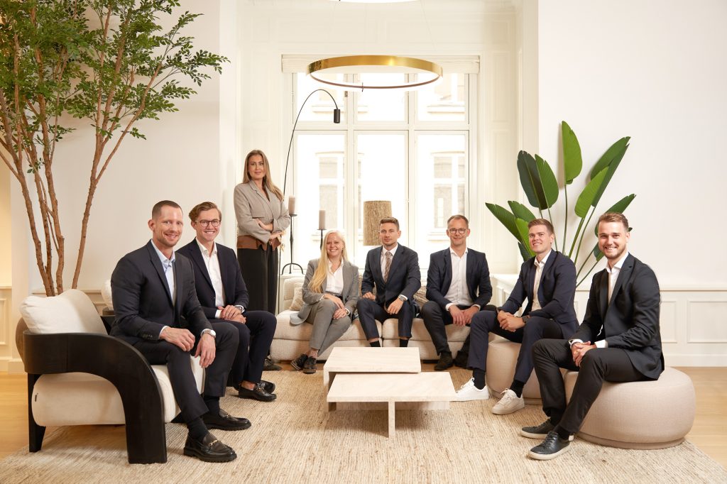 Otte kolleger i lyst erhvervskontor med sofa og grønne planter, afslappet gruppefoto til virksomhedshjemmeside