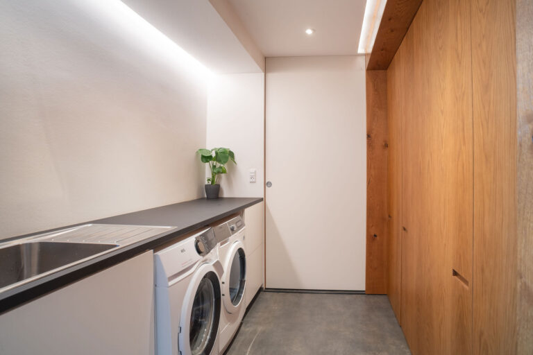 Moderne vaskerum, der er perfekt til en Airbnb, med vaskemaskine og tørretumbler under en elegant sort bordplade, en vask til venstre, træskabe til højre, en potteplante, der giver friskhed, og en hvid skydedør på bagsiden - ideelt til enhver fotograf.