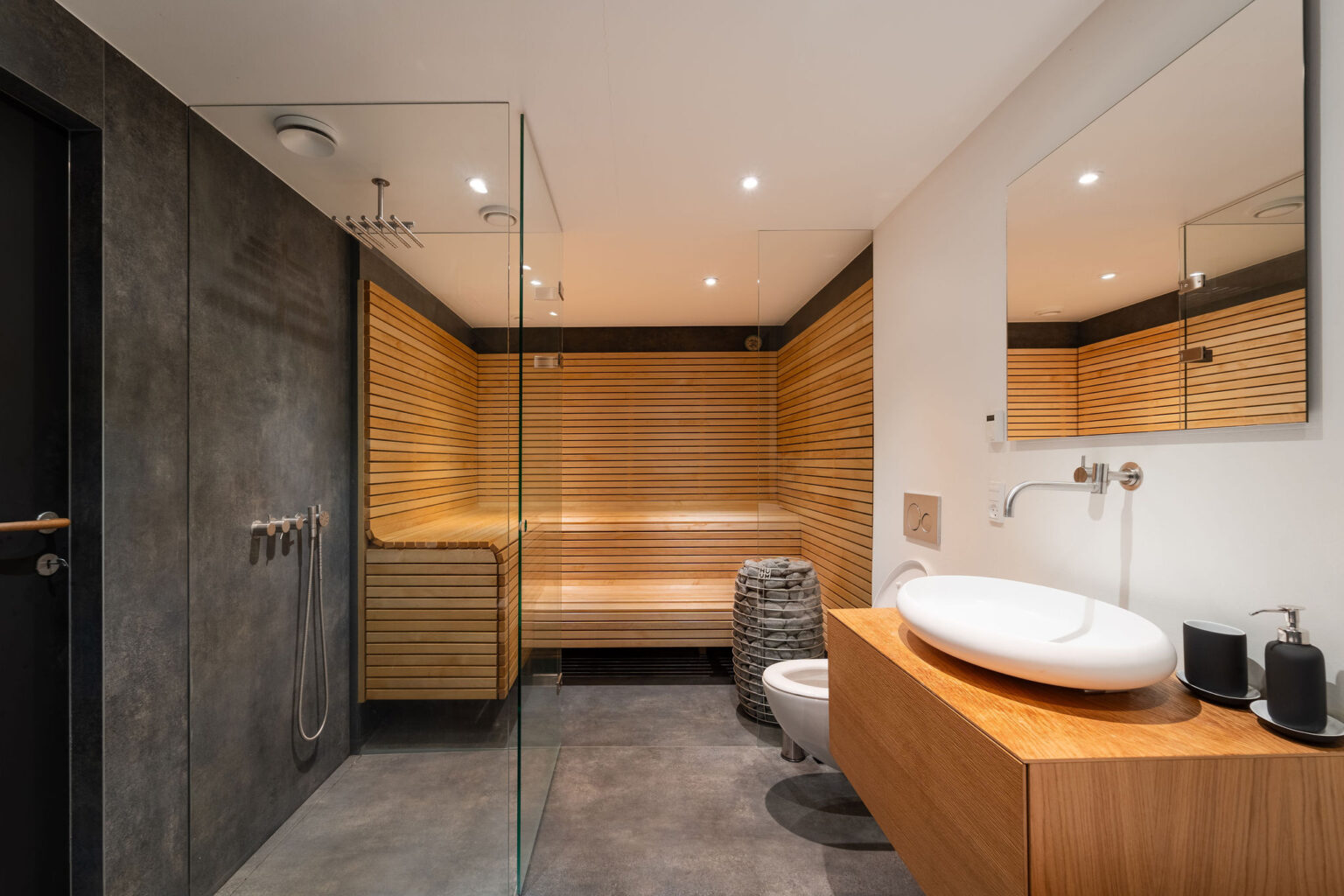 Det moderne badeværelse i denne Airbnb har en glasindkapslet bruser, sauna i træ, væghængt toilet og håndvask i træ med kar; perfekt til fotoentusiaster, der sætter pris på minimalistisk design og varm belysning.