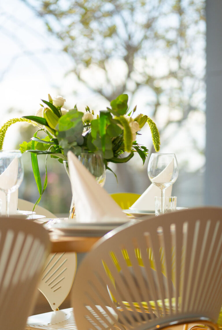 Et nærbillede af et spisebord dækket til en Airbnb, med hvide servietter, vinglas og en grøn blomsterdekoration, fanget af en fotograf i blødt naturligt lys med slørede træer, der kan ses gennem vinduet.