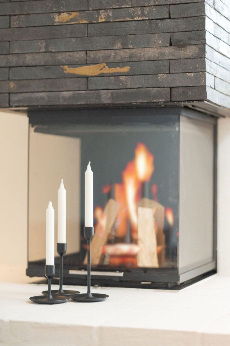 Tre hvide stearinlys i sorte lysestager sidder foran en moderne, glasindkapslet pejs, hvor ilden gløder varmt. Disse hyggelige, minimalistiske omgivelser er perfekte for en Airbnb-fotograf til at tage stilfulde og indbydende interiørbilleder.
