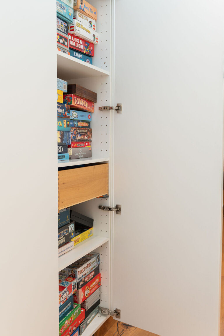 Et hvidt skab med flere hylder er fyldt med stablede brætspil, perfekt til en Airbnb-spilaften. Skabslågen er delvist åben og afslører farverige kasser, mens en træskuffe er trukket lidt ud - ideel til fotografisk inspiration.