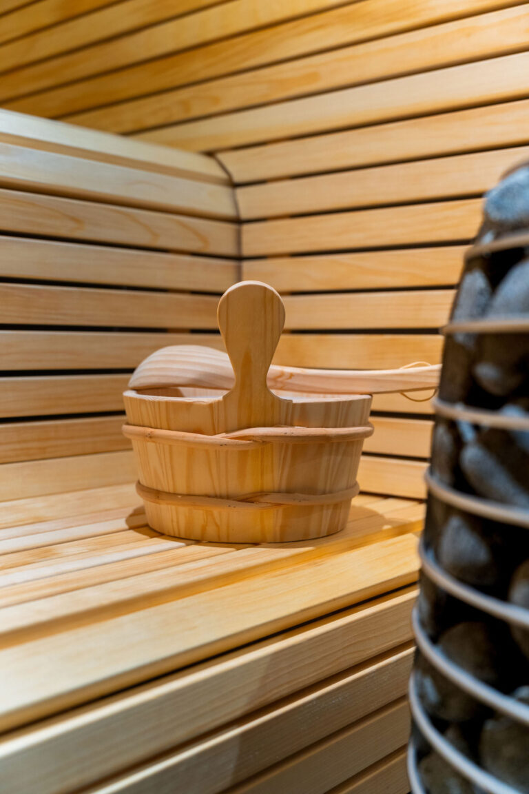 En træspand med en øse står på en bænk inde i en Airbnb-sauna med træpaneler på væggene og siddepladserne, og en del af saunaovnen, som fotografen har fanget, ses til højre.
