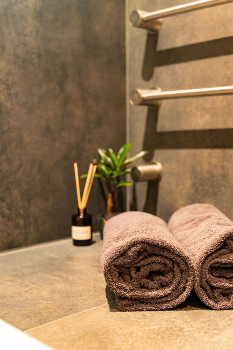 To sammenrullede grå håndklæder hviler på en flisebelagt badeværelsesoverflade nær en lille potteplante og en diffuser, hvilket skaber en fotografisk Airbnb-stemning med slanke håndklædestænger i metal monteret på væggen i baggrunden.