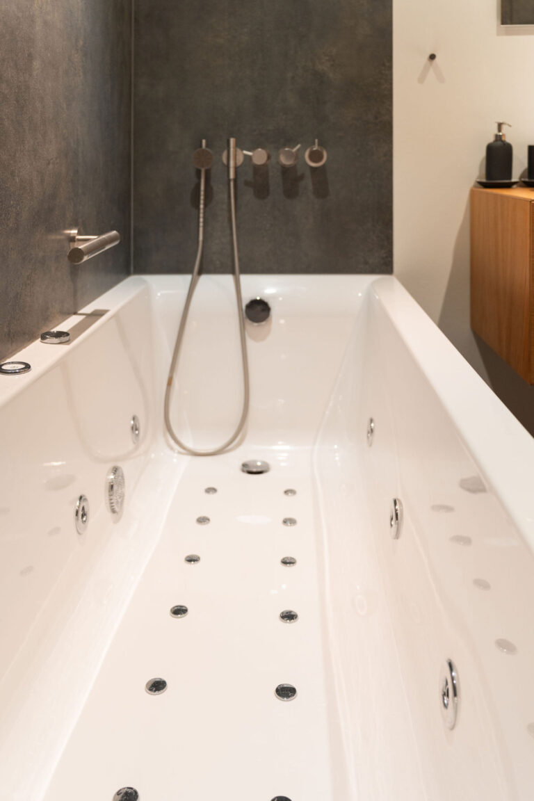 Moderne hvidt boblebad med flere stråler, et håndholdt brusehoved og minimalistiske armaturer mod en mørk struktureret væg - perfekt til Airbnb-opslag eller fotografering; sæbedispenser og skab er synlige på siden.