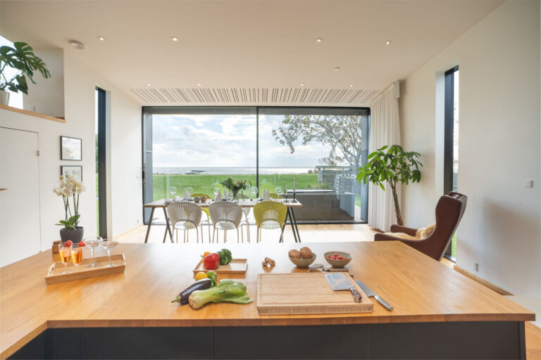 Moderne køkken og spiseplads, perfekt til Airbnb-ophold, med store glasdøre ud til en grøn græsplæne. Grøntsager og køkkenudstyr står på en træø, mens sollyset lyser op i dette rummelige, minimalistiske rum - ideelt for enhver fotograf, der søger inspiration.