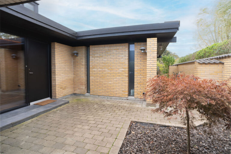 Moderne hus udvendigt med gule murstensvægge, sort kant og en sort hoveddør. Det er ideelt til Airbnb-ophold og har en brolagt gårdhave og et anlagt bed med et rødligt træ - en perfekt scene for enhver fotograf.