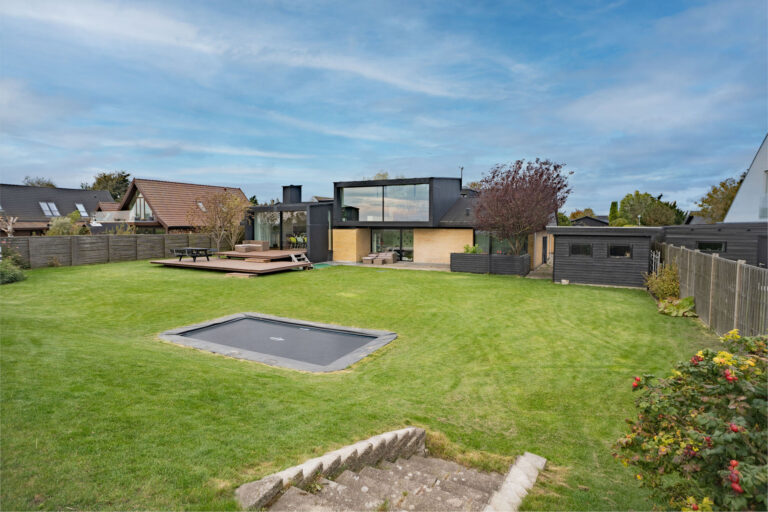 Moderne hus med store vinduer og en rummelig baghave, perfekt til Airbnb-fotos, med en nedsænket trampolin og træterrasse. Den indhegnede have er omkranset af træer og buske og ligger under en delvis overskyet himmel.