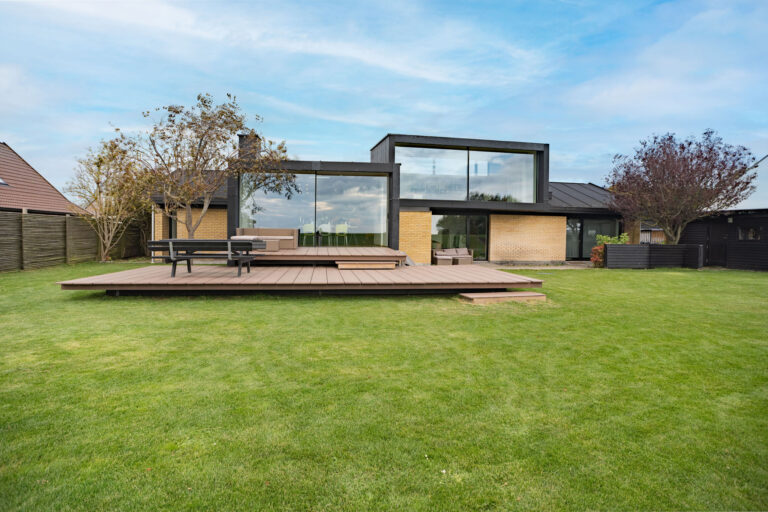 Moderne hus med store glasvinduer og en rummelig træterrasse i en græsklædt baghave - perfekt til dit næste Airbnb-ophold. Billedet er taget af en dygtig fotograf og har frodige træer og en blå himmel i baggrunden.