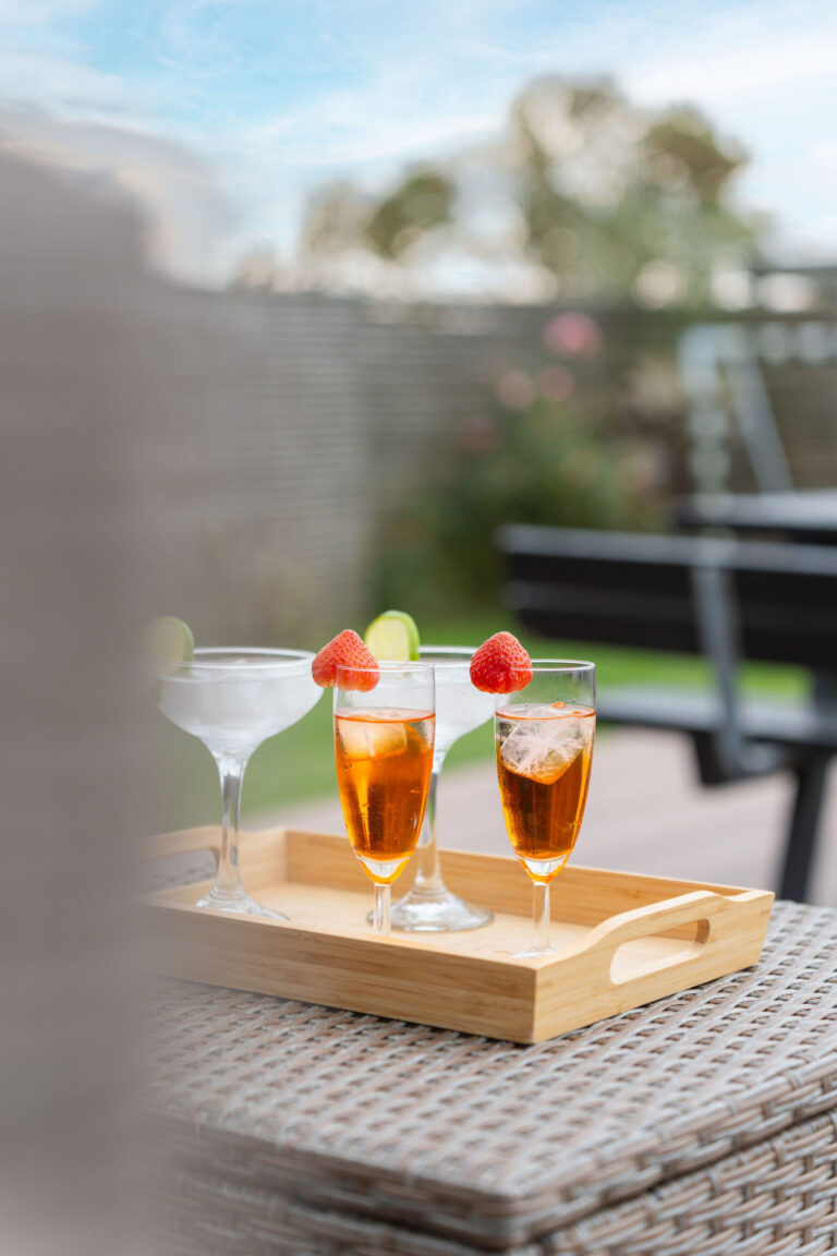 En træbakke på et flettet bord indeholder to cocktails pyntet med jordbær og en klar drink med limebåd - et indbydende Airbnb-miljø fanget af en dygtig fotograf med slørede grønne planter og en bænk i baggrunden.