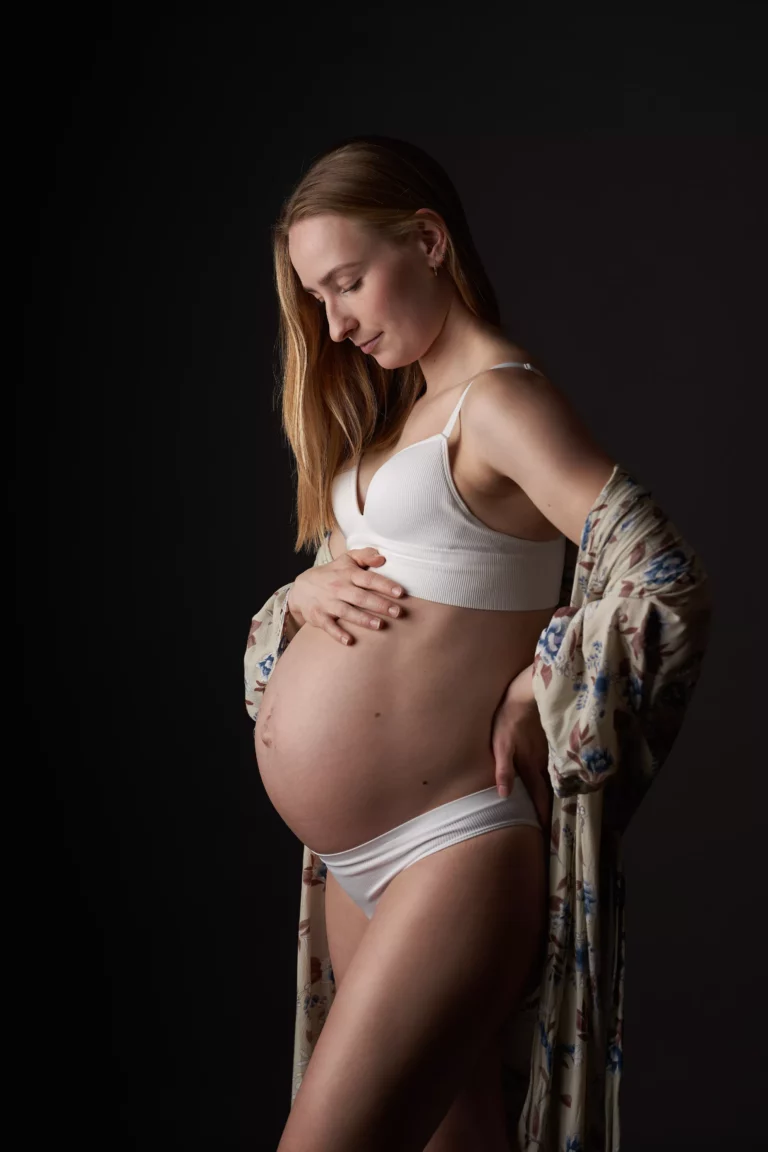En gravid kvinde står i profil mod en mørk baggrund, iført hvid bh og undertøj med en blomstret kåbe over armene, mens hun forsigtigt vugger sin mave og kigger ned under et Gravid fotosession-studie.