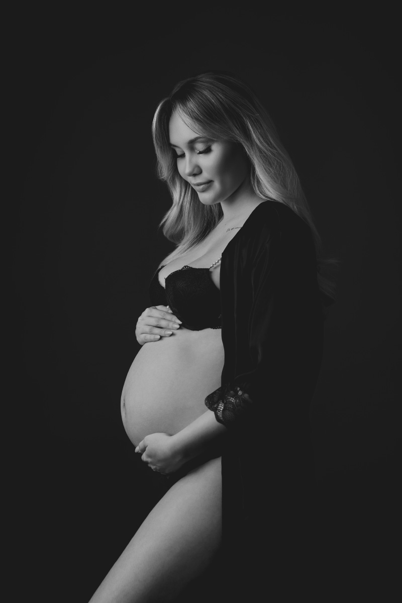 Sort-hvidt foto fra en Gravid-fotosession i et fotostudie, der viser en gravid kvinde i lingeri og morgenkåbe, som forsigtigt holder om sin mave med begge hænder og ser fredeligt ned mod en mørk baggrund. Pris 899 kr.