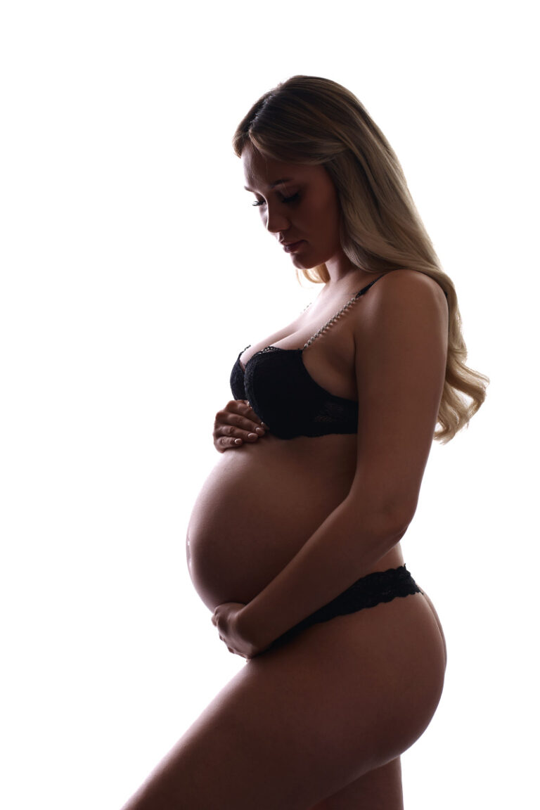 En gravid kvinde i sort lingeri ses i profil mod en hvid baggrund, mens hun forsigtigt holder om sin mave med den ene hånd - et fredfyldt øjeblik fra en gravid fotosession af Weigang Fotografi, og hendes eftertænksomme udtryk er fuld af forventning.