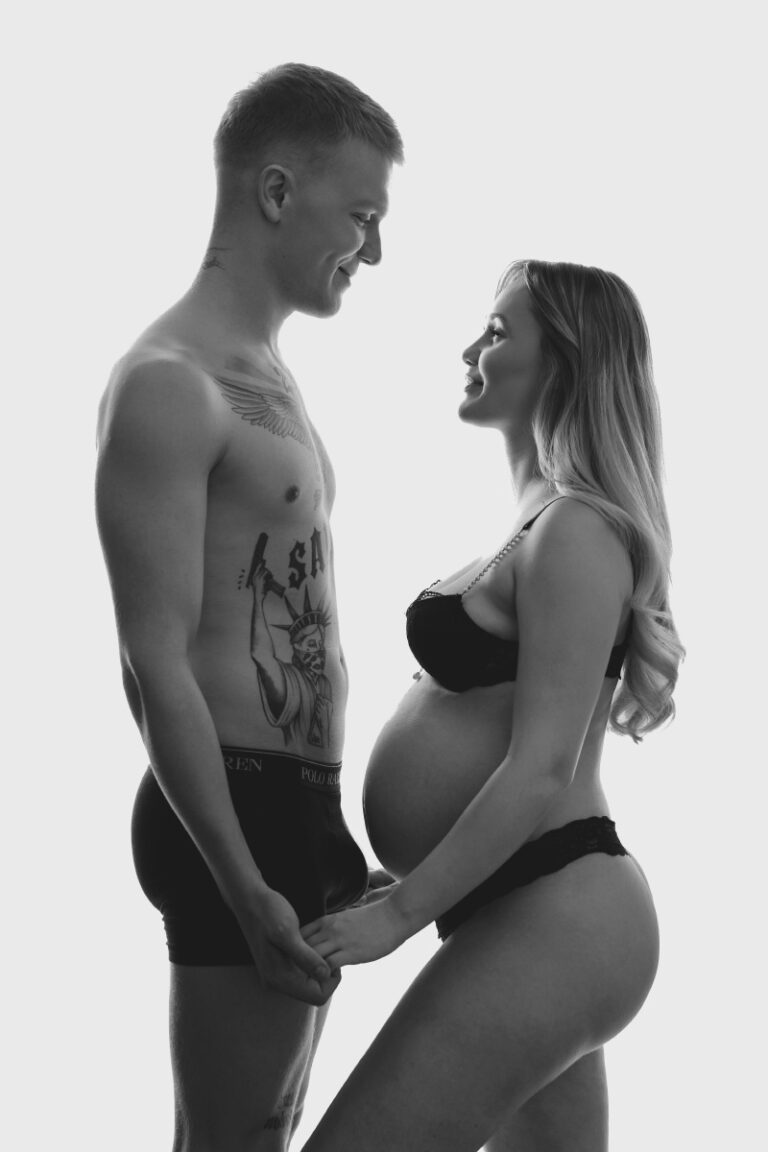 En smilende mand og en gravid kvinde i undertøj står over for hinanden, holder hinanden i hånden og kigger hinanden i øjnene mod en lys baggrund.