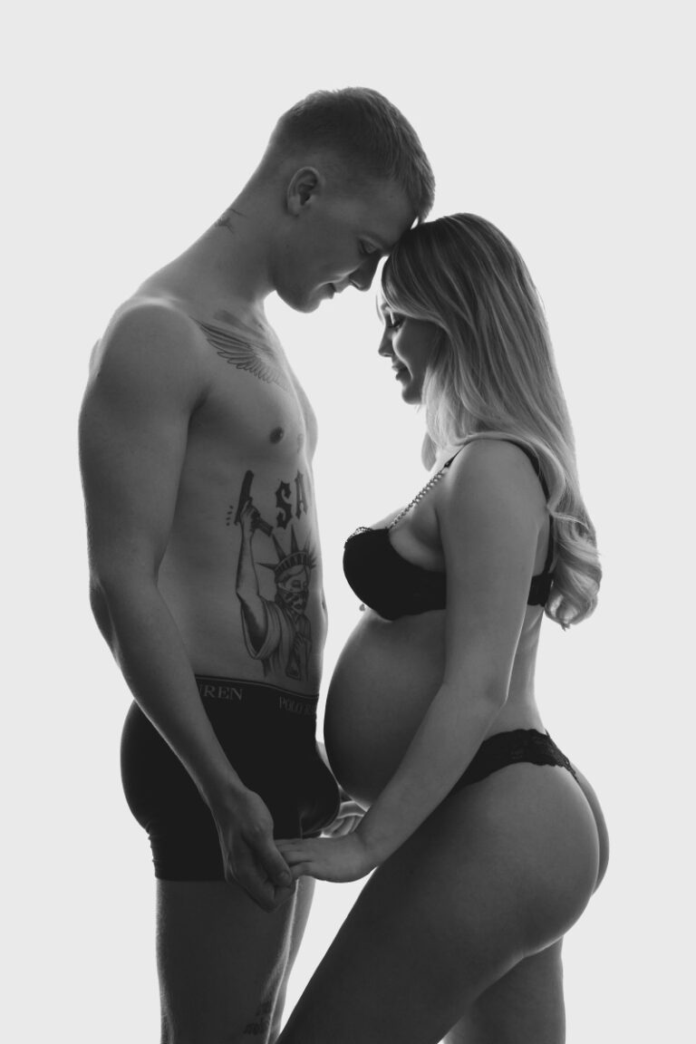 En mand i bar overkrop og en gravid kvinde i sort lingeri står over for hinanden, holder hinanden i hånden og rører ved hinandens pander under en Weigang Fotografi gravid fotosession med en hvid baggrund bag dem.