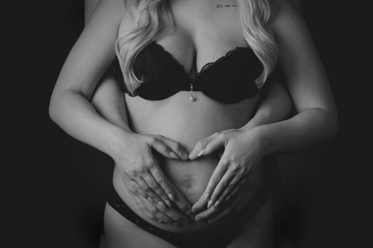 En gravid kvinde i sort lingeri bliver omfavnet bagfra under en gravid fotosession. Både hendes og hendes partners hænder hviler på hendes bare mave og danner en hjerteform mod den mørke baggrund, som fremhæver parret og babybulen.