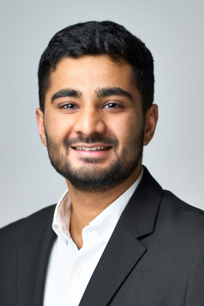 LinkedIn-portræt af ung mand med kort skæg i sort jakkesæt og hvid skjorte, bredt og varmt smil, lys grå studiobaggrund