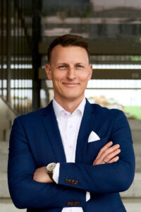 Professionelt portræt af en mand iført en blå jakkesæt og hvid skjorte, taget udendørs med moderne arkitektur i baggrunden. Ideelt til LinkedIn og erhvervsprofiler. Erhvervsfotografering fra Weigang Fotografi i København.