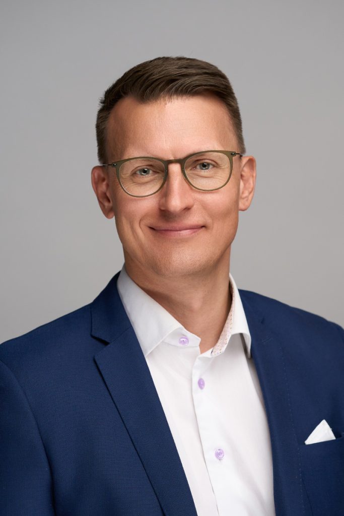 LinkedIn-portræt af mand med briller i blåt jakkesæt og hvid skjorte, roligt venligt smil mod grå studiobaggrund