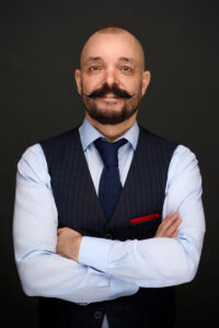 En mand med skaldet hoved og stylet overskæg, iført lyseblå skjorte, marineblå vest og slips, står med korslagte arme mod en mørk baggrund - et professionelt linkedin-billede fanget af en erfaren erhvervsfotograf. En rød lommeklud fremhæver hans vest.