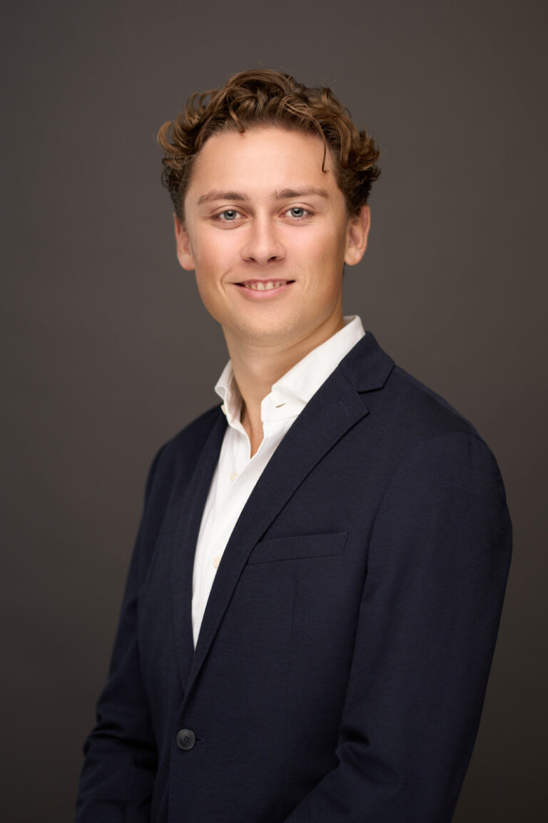 Professionelt portræt af en ung mand iført en mørk blazer og hvid skjorte, taget i et studie med en neutral, mørk baggrund.