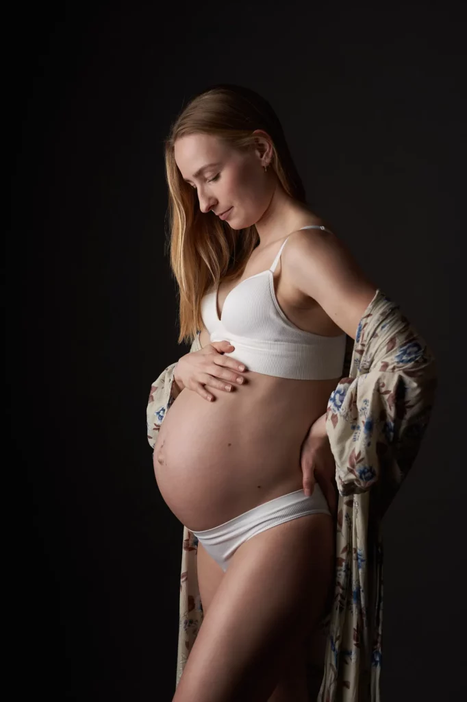 Pregnant woman in white underwear and flower kimono looks down at her belly and holds it with both hands against a dark background