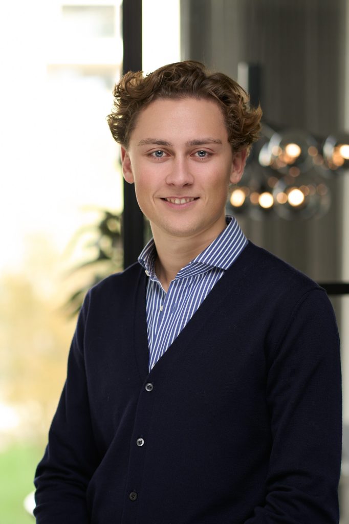 Ung mand med krøllet hår i marineblå sweater over stribet skjorte, headshot til medarbejderprofil