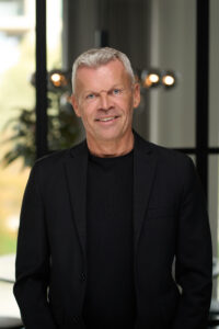 En smilende ældre mand med kort gråt hår iført sort blazer og sort skjorte står indendørs foran et vindue med sløret lys og en plante, fanget i en professionel forretningsportræt-stil.