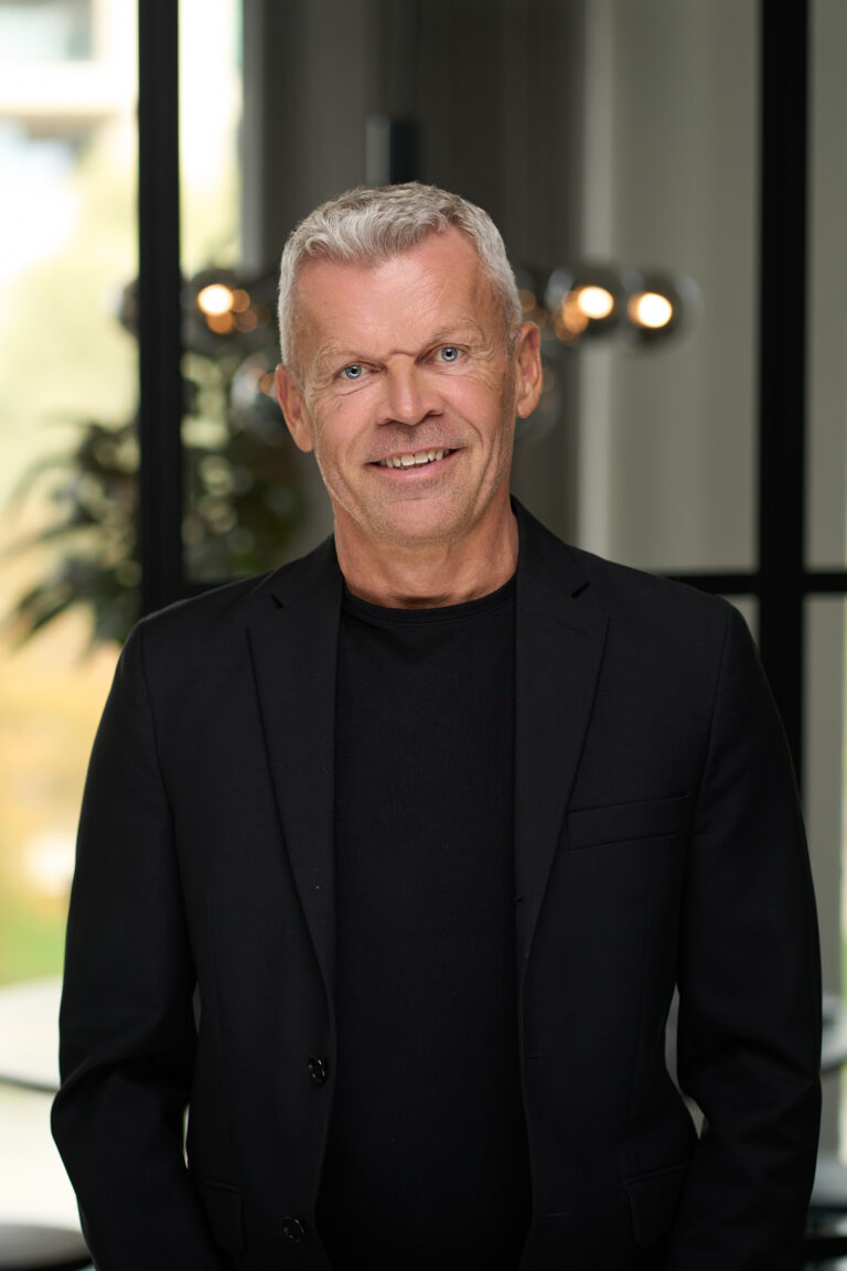En smilende ældre mand med kort gråt hår iført sort blazer og sort skjorte står indendørs foran et vindue med sløret lys og en plante, fanget i en professionel forretningsportræt-stil.