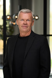 En moden mand med kort gråt hår iført sort blazer og sort skjorte står indendørs og smiler let i et sløret moderne miljø - et ideelt forretningsportræt, der indfanger professionalisme.