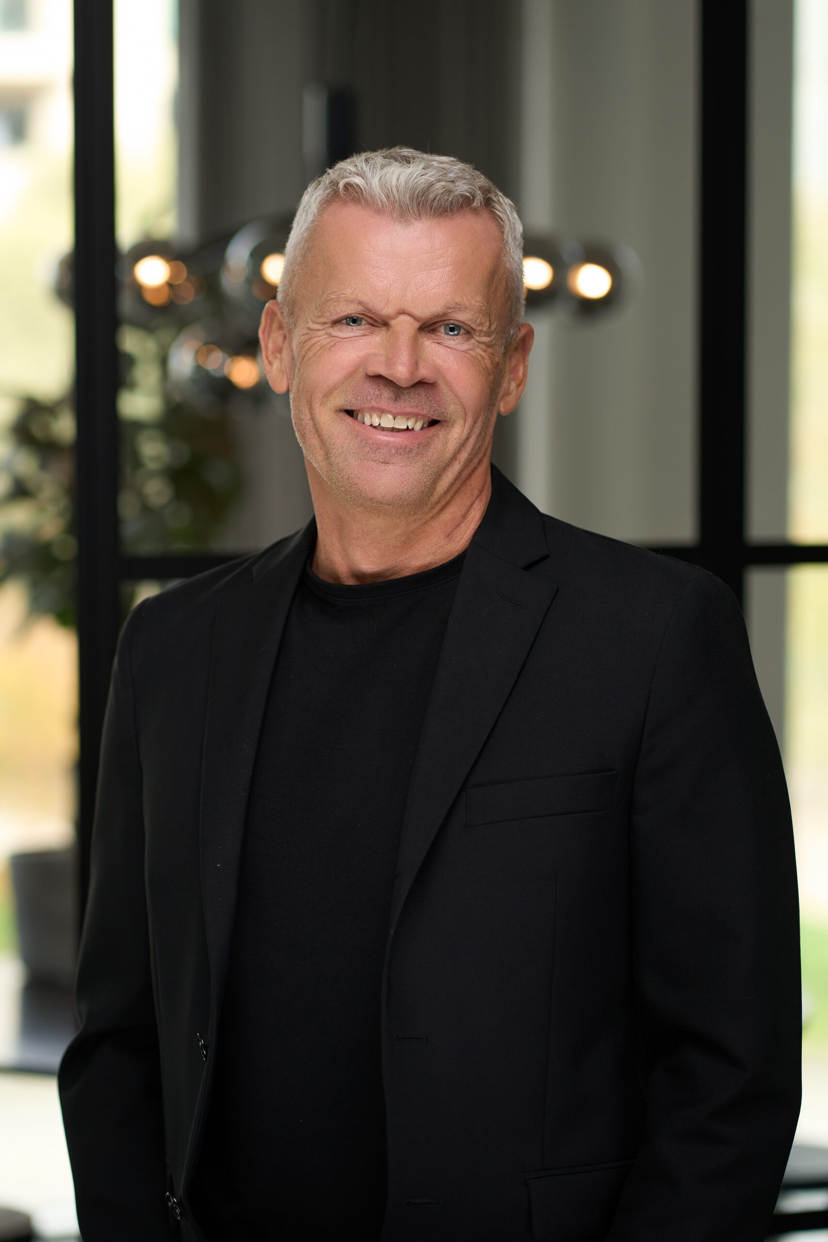 En smilende midaldrende mand med kort gråt hår iført sort blazer og sort skjorte står indendørs i et moderne, veloplyst rum med sløret baggrund, perfekt til et forretningsportræt eller et medarbejderportræt.