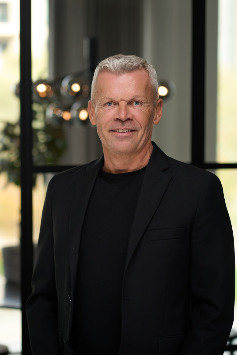 Professionelt portræt af en mand iført en sort blazer og sort t-shirt, taget i et moderne kontormiljø med slørede detaljer af glasvægge og belysning i baggrunden.