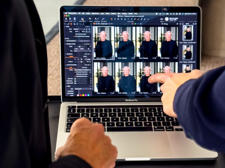 To personer gennemgår medarbejderportræt-headshots på en MacBook Pro. Den ene peger på skærmen, der viser forskellige billeder af en mand i en sort skjorte, mens den anden observerer. Et fotoredigeringsprogram er åbent på den bærbare computer.