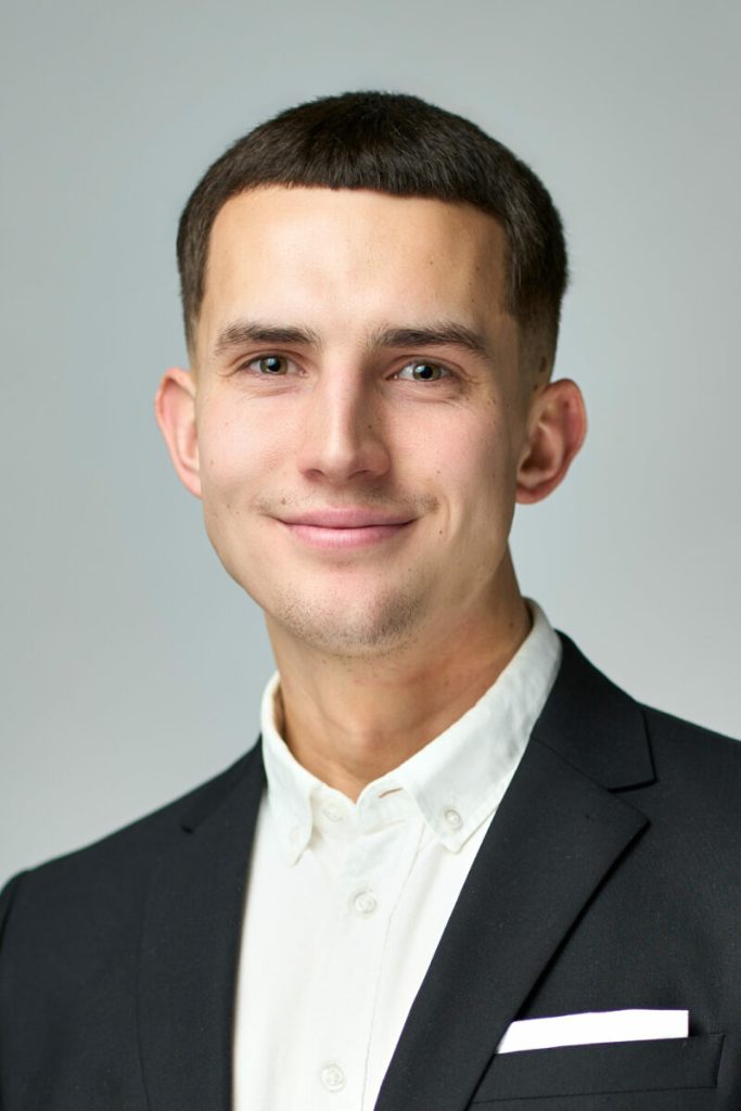 Ung mand med kort mørkt hår i sort blazer og hvid skjorte, portræt mod grå studiobaggrund, medarbejderfoto