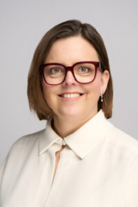 En kvinde med kort brunt hår, iført store røde briller, en cremefarvet bluse med krave og øreringe, smiler til kameraet mod en almindelig lys baggrund - et ideelt LinkedIn-billede.