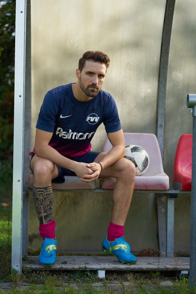 Fodboldspiller i blå og pink Nike-trøje sidder på reservebænken med en fodbold, sportportræt udendørs