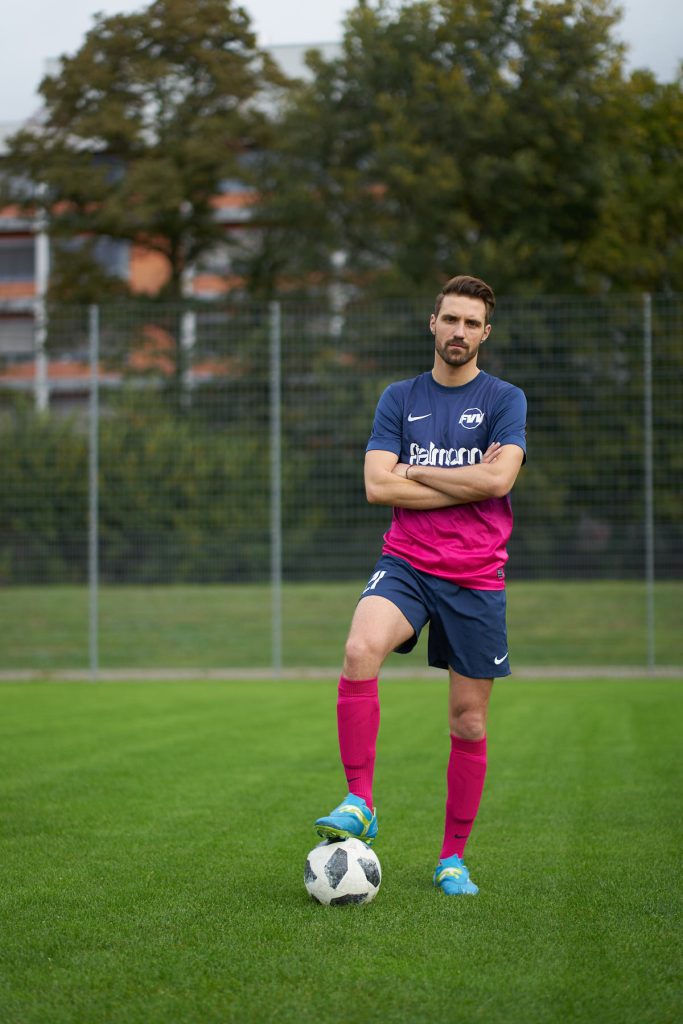 Fodboldspiller i navyblå og pink trøje med foden på bold og armene over kors på grøn fodboldbane