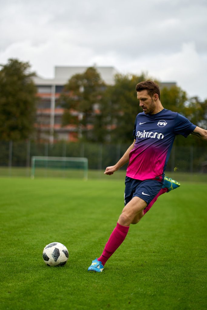 Fodboldspiller i navy og pink trøje tager et spark til bold på grøn fodboldbane, aktionsbillede