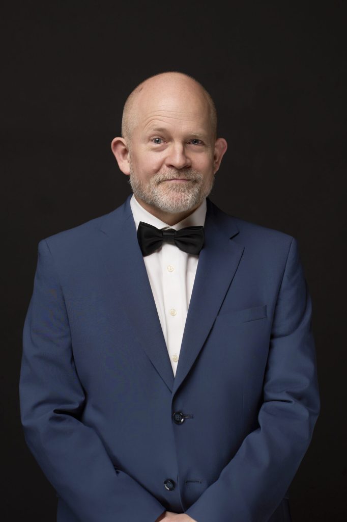 Skaldet mand med gråt skæg i blå jakkesæt og sort butterfly mod sort baggrund, studioportræt