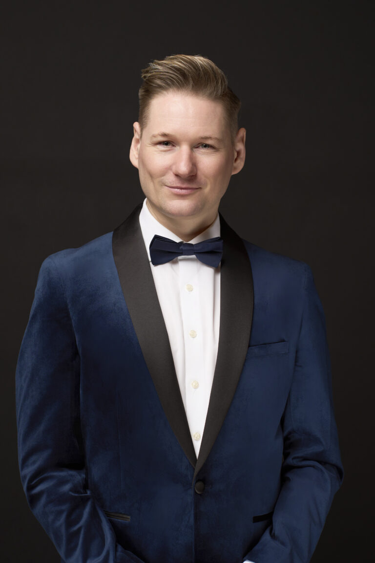 Portræt af skuespiller: En mand med kort, stylet hår iført blå fløjlsjakke med sort revers, hvid skjorte og sort butterfly står på en ensfarvet, mørk baggrund og smiler let til kameraet.