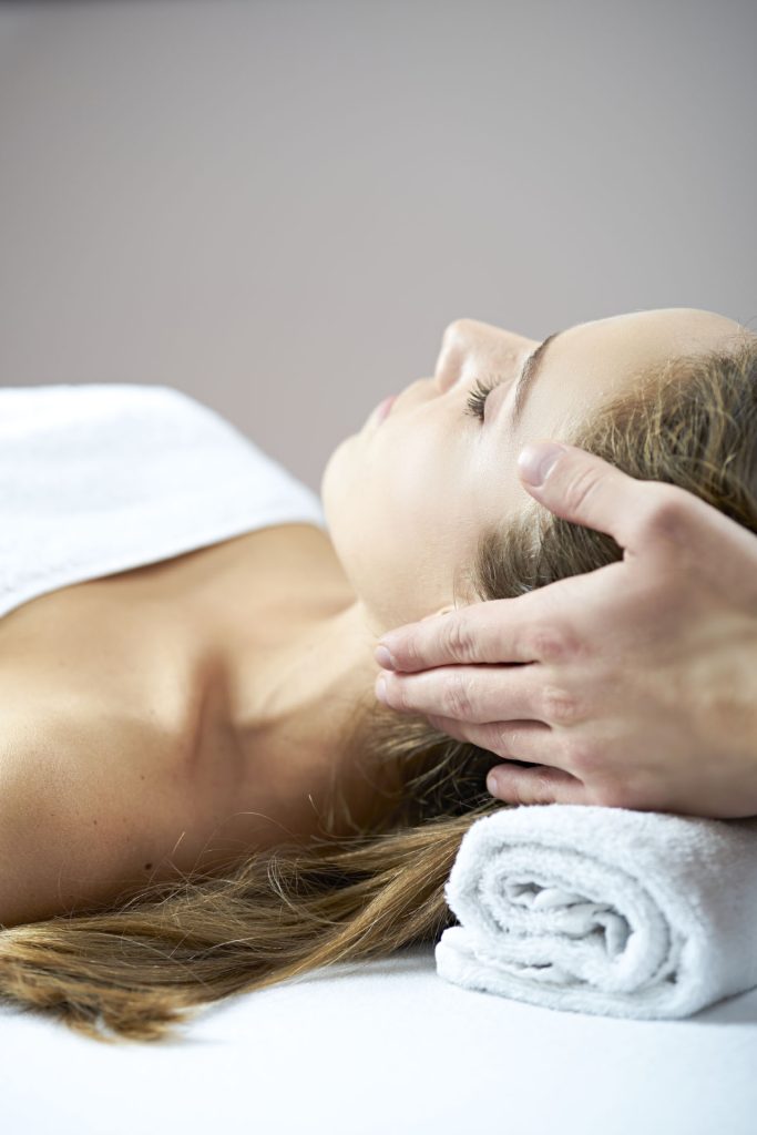 Kvinde modtager kranio-massage på behandlingsbrik, ansigt vendt opad med lukkede øjne og håndklæde