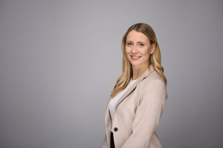 En kvinde med langt blondt hår iført beige blazer og hvid top smiler til kameraet i dette elegante portræt på en almindelig lysegrå baggrund.