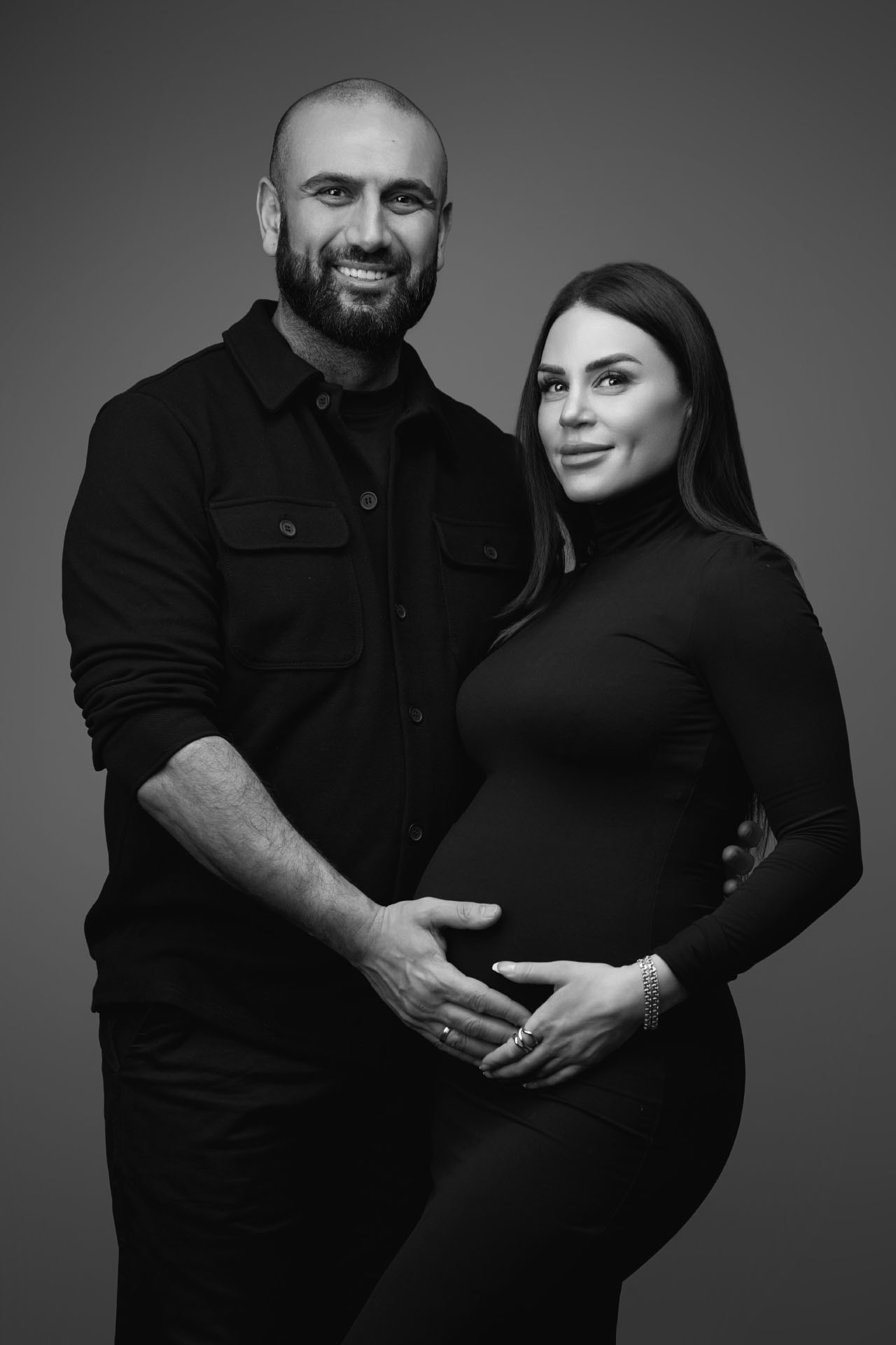 Sort-hvid graviditetsfoto af skaldet mand med skæg og gravid kvinde i sort turtleneck der begge smiler mod kameraet