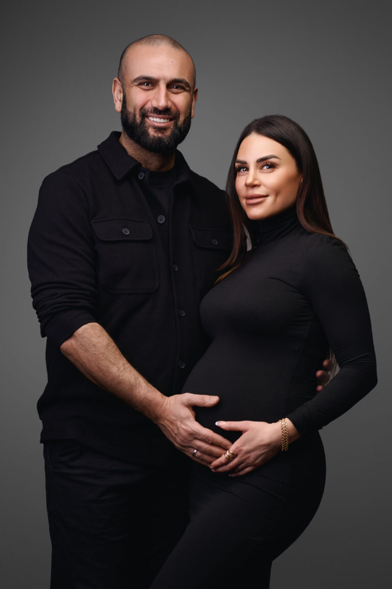 Et smilende par poserer sammen i en gravid fotosession, begge klædt i sort. Manden hviler blidt sine hænder på kvindens gravide mave, mens hun smiler blidt og holder hans hænder. Baggrunden er enkel og mørk og skaber et klassisk studie-look.