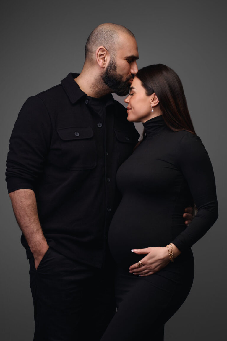 En mand kysser en gravid kvindes pande under deres Gravid-fotosession. Begge er klædt i sort, hun holder sin babybule, og de står tæt sammen og ser fredfyldte ud mod en mørkegrå studiebaggrund.