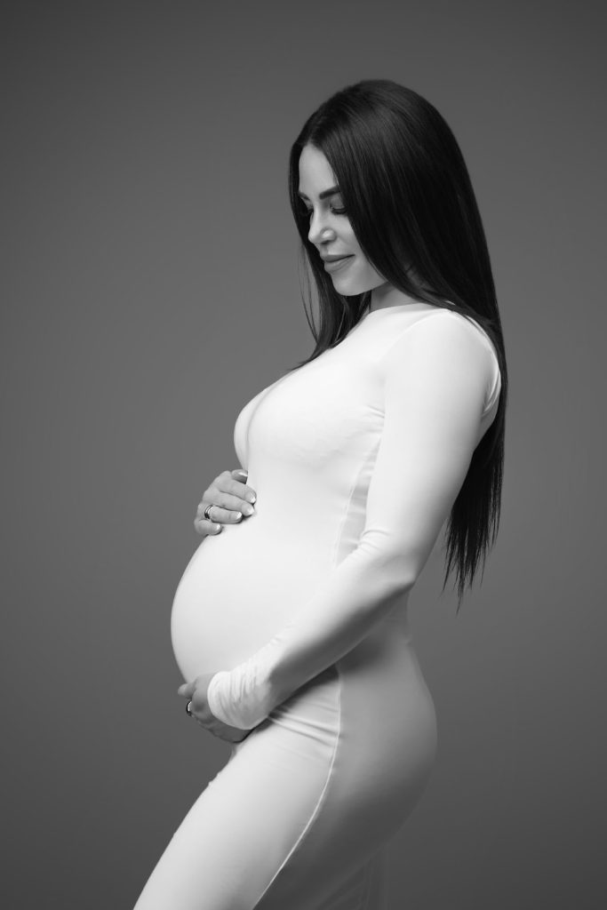 Sort-hvid graviditetsfoto af kvinde i hvid tætsiddende bodysuit med langt sort hår i profil mod grå baggrund